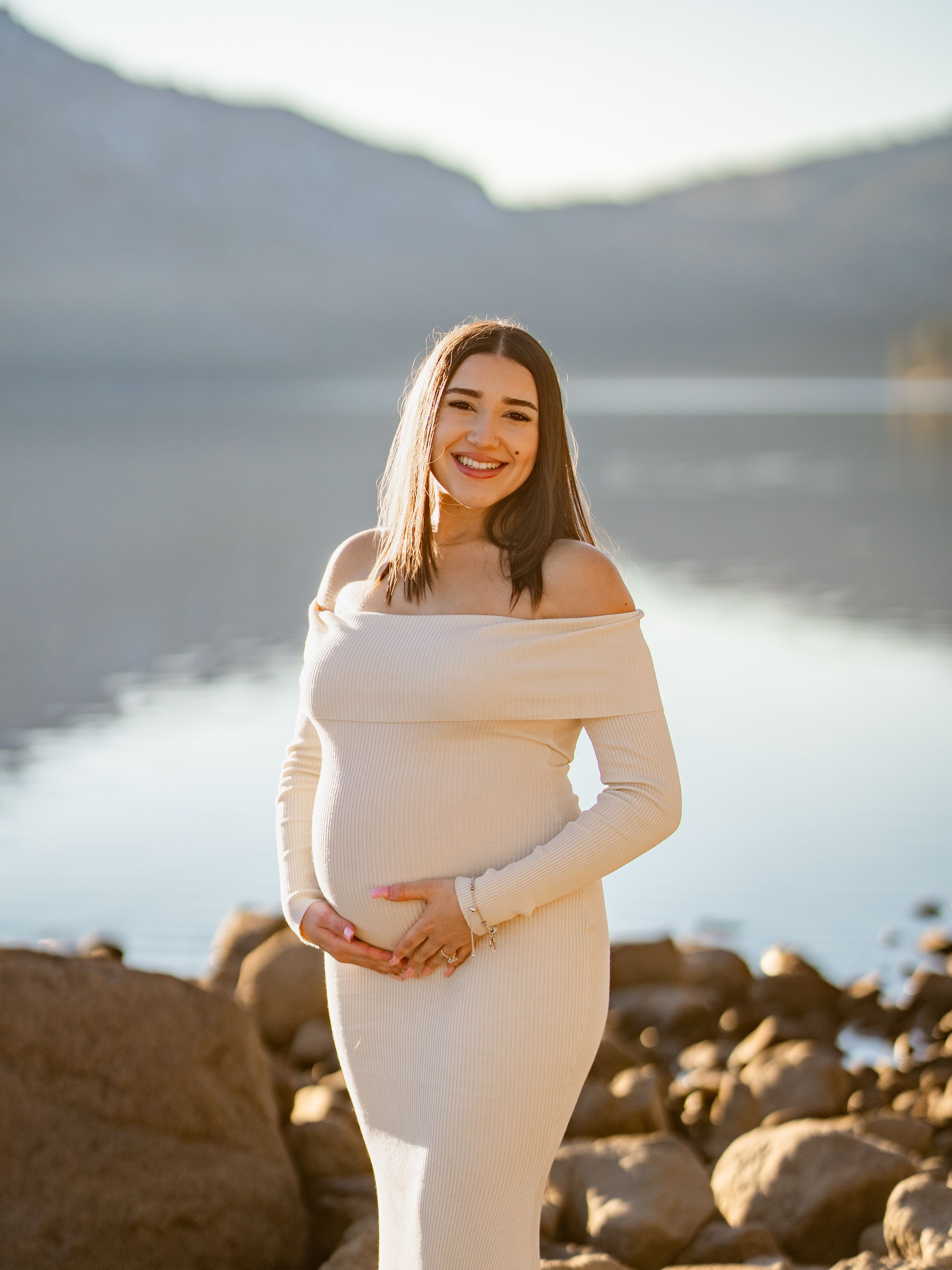 Expectant mother in white dress poses at lakeside during golden hour with mountains in background.