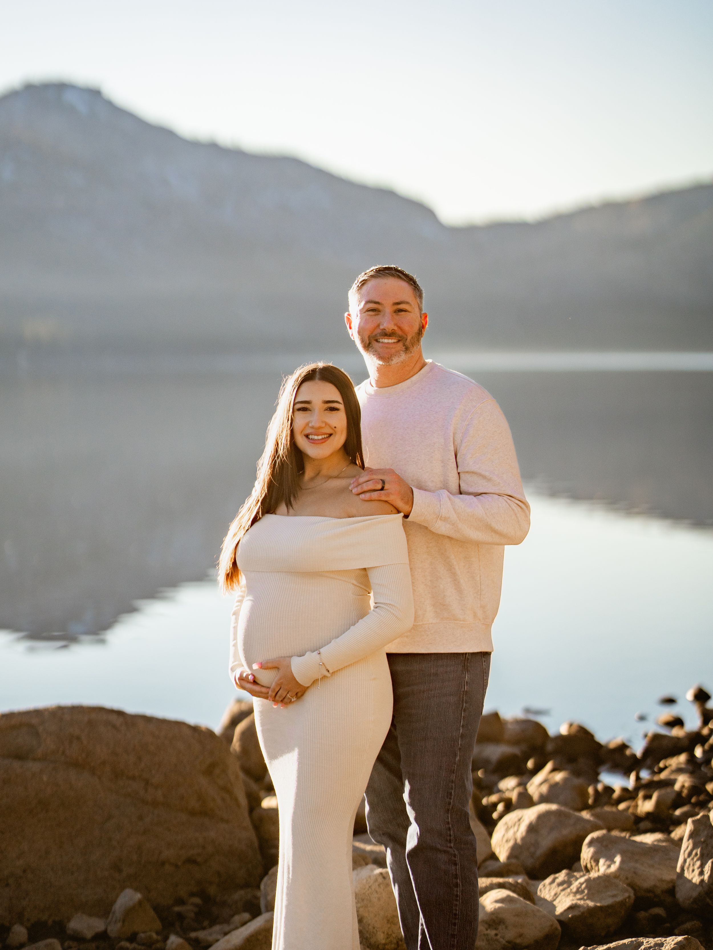 Romantic maternity photography session at sunset by a serene mountain lake with rocky shoreline.