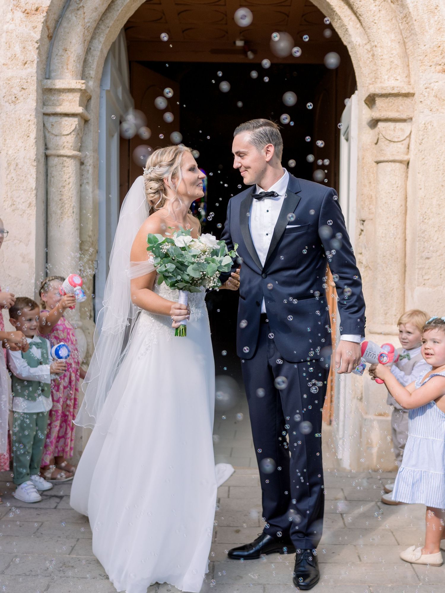 Newlyweds celebrate under stone archway as bubbles float through the air.