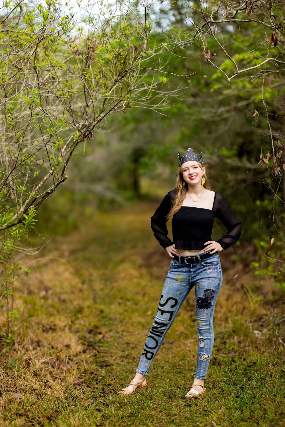 A person wearing ripped jeans and a black top poses casually among green bushes and foliage in an outdoor setting.