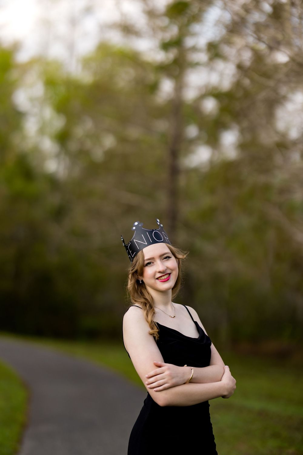 A graduate in a black dress and cap poses outdoors in a natural setting with trees in the background.