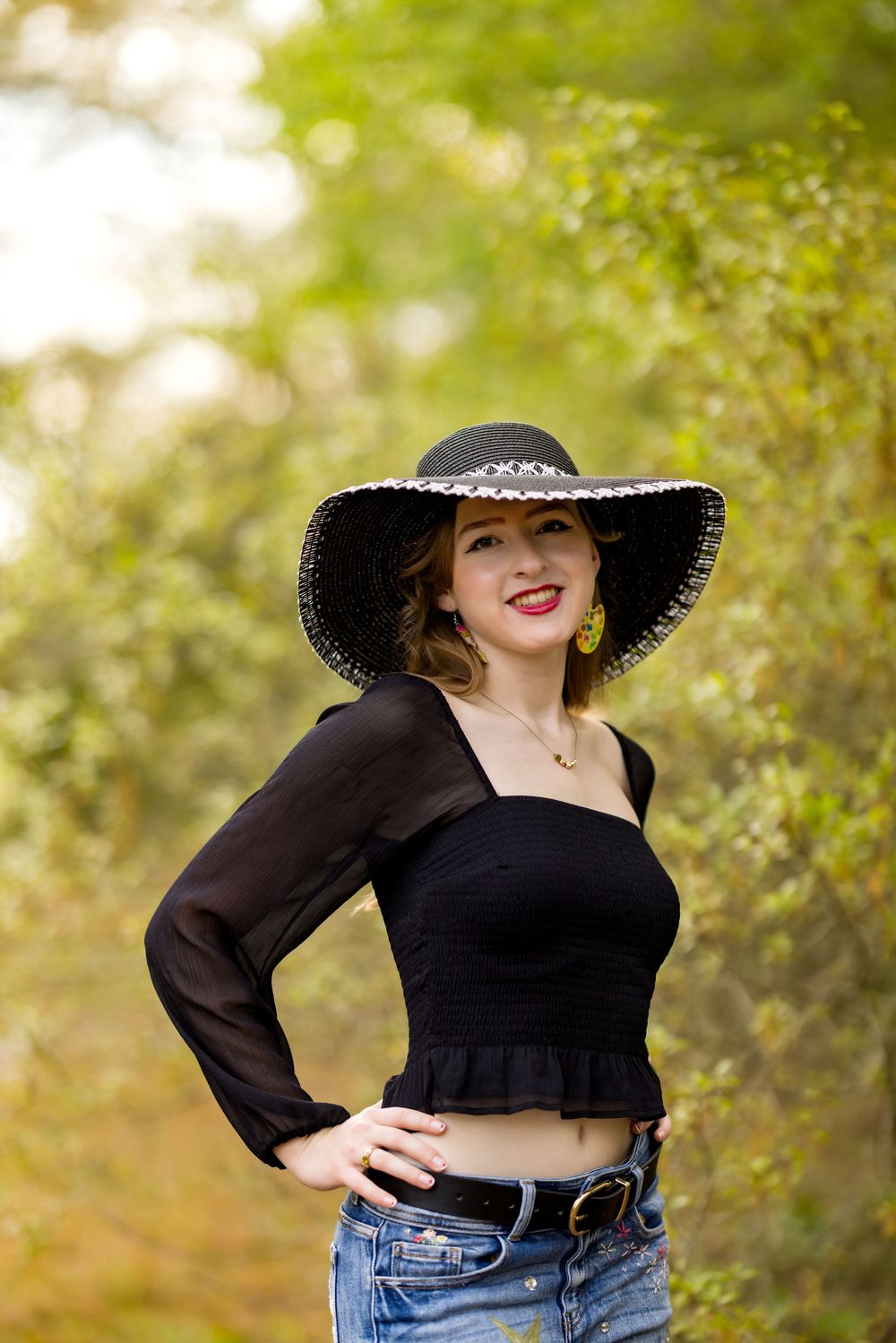 A person in a wide-brimmed black hat and cropped top poses outdoors against a blurred natural background.