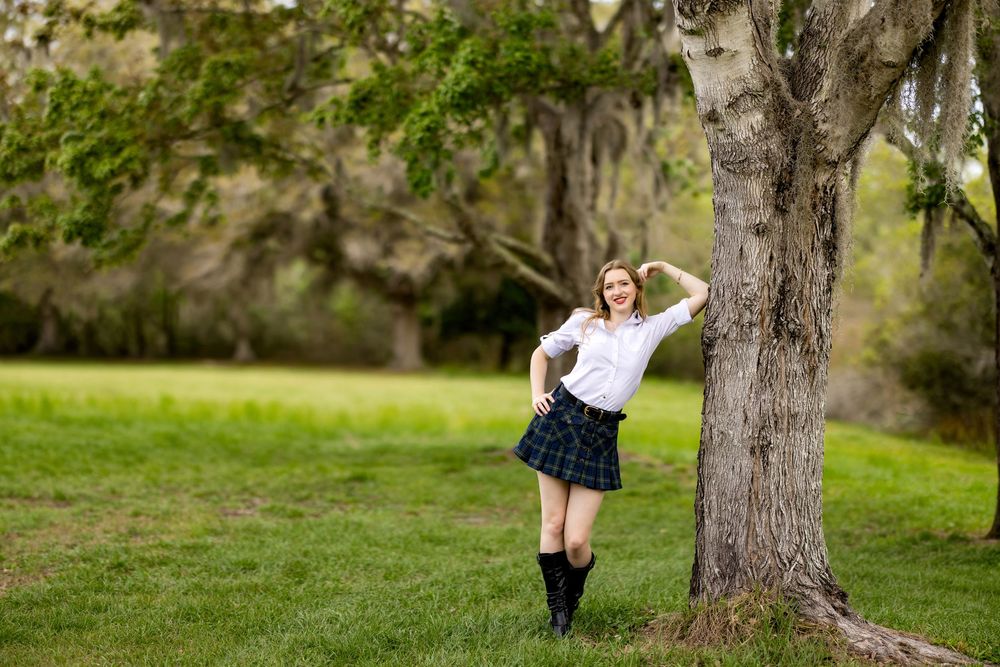 A person in a white sweater and navy skirt stands next to a tree in a grassy park setting on a sunny day.