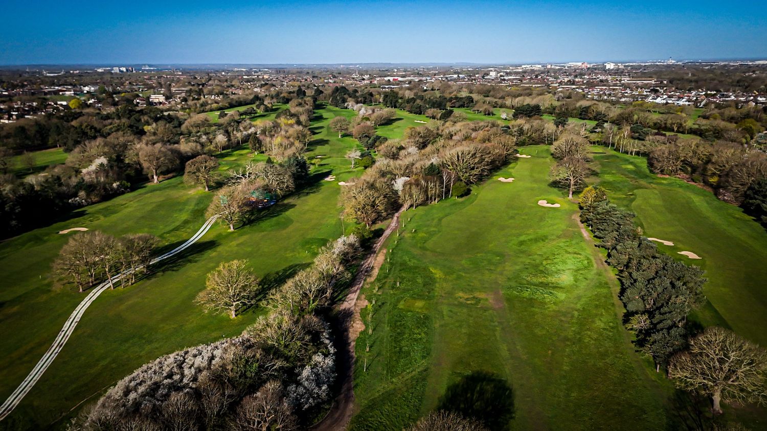 Aerial view of a lush green golf course with winding fairways and tree-lined paths under bright blue sky.