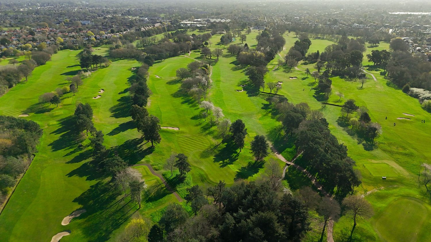 Aerial panoramic view of a lush green golf course with trees and residential areas in the background.