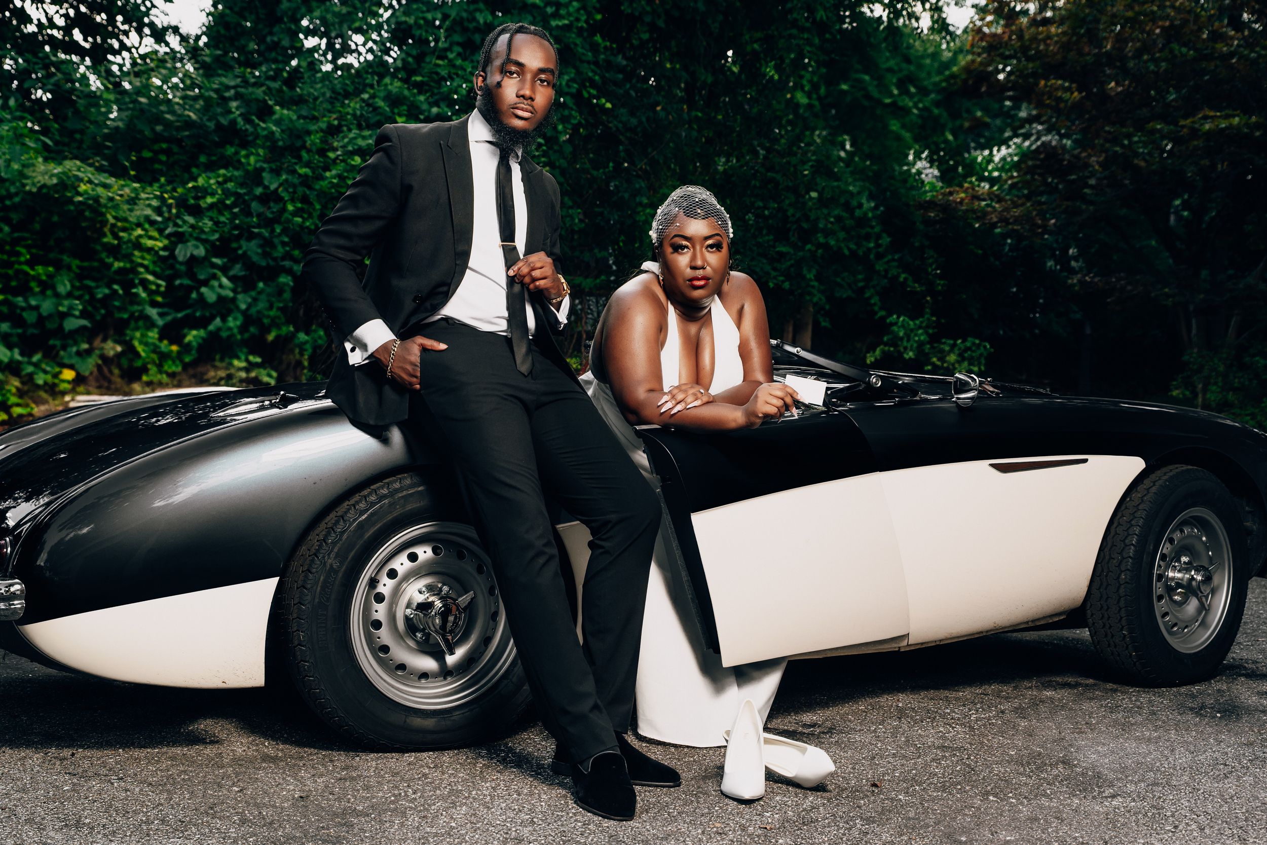 A couple poses with a classic black and white vintage sports car on a tree-lined driveway.