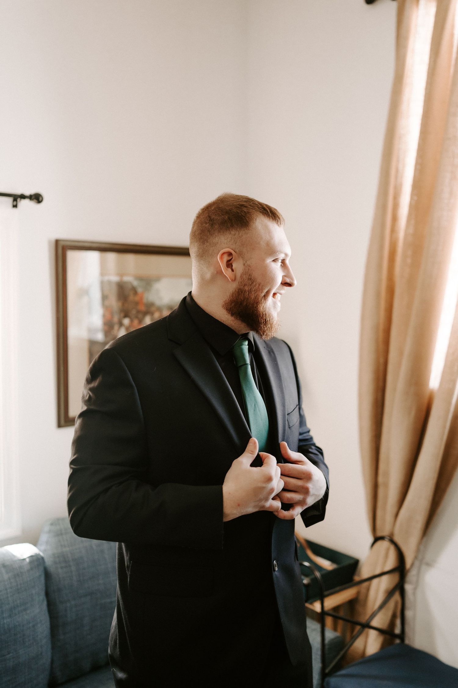 Dapper red-haired groom adjusting black suit with green tie near window.