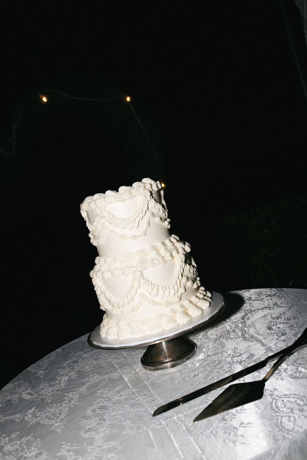 Elegant three-tier white wedding cake with ruffled frosting on a silver stand, displayed outdoors at night.