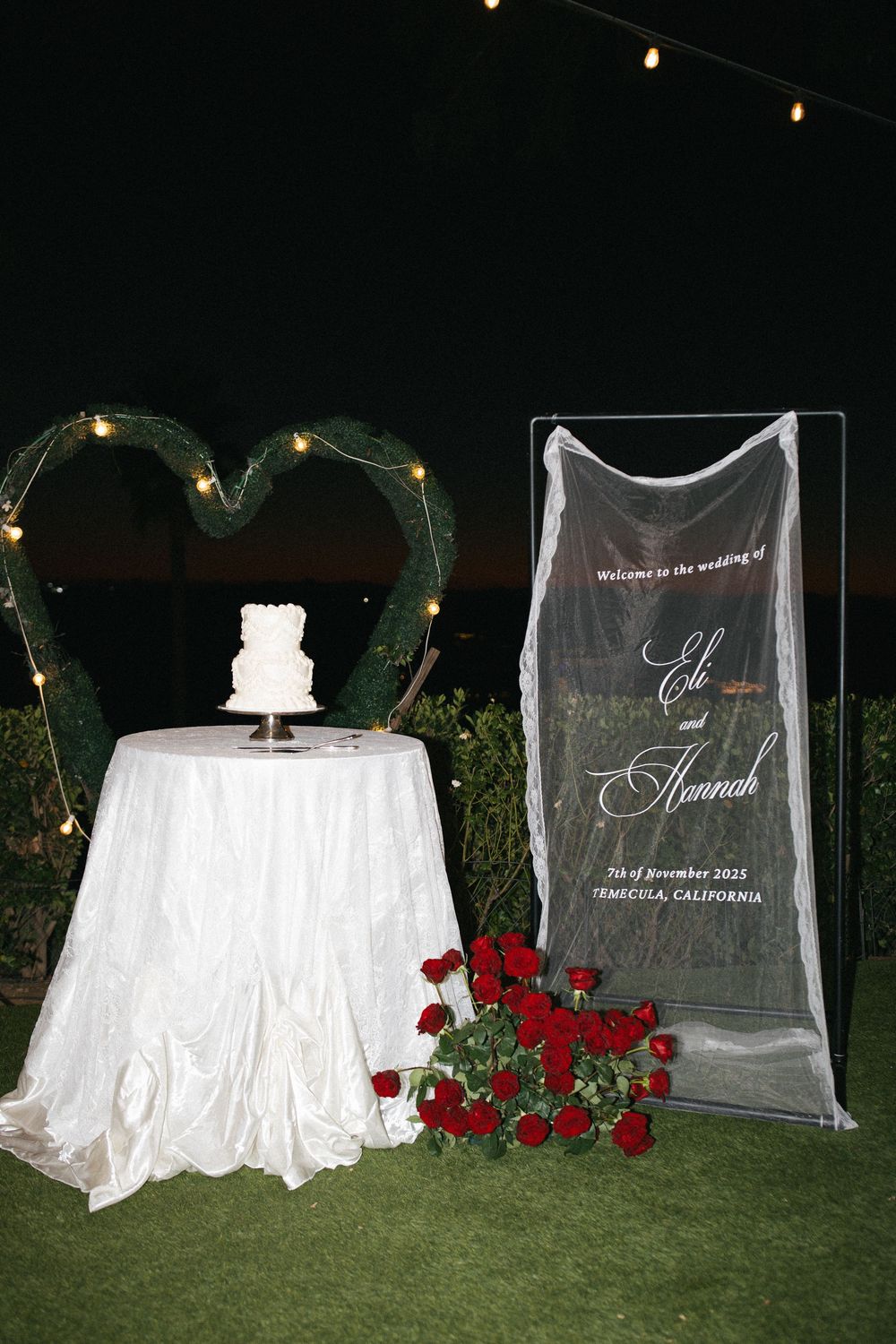 Elegant outdoor wedding display with white cake, heart-shaped greenery arch, red roses, and acrylic welcome sign in Temecula, CA.