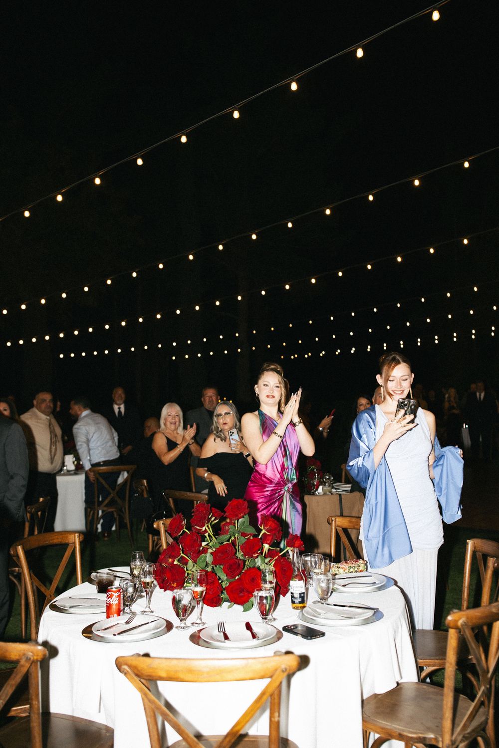 Guests in elegant attire applaud at an outdoor wedding reception with string lights and red rose centerpieces.