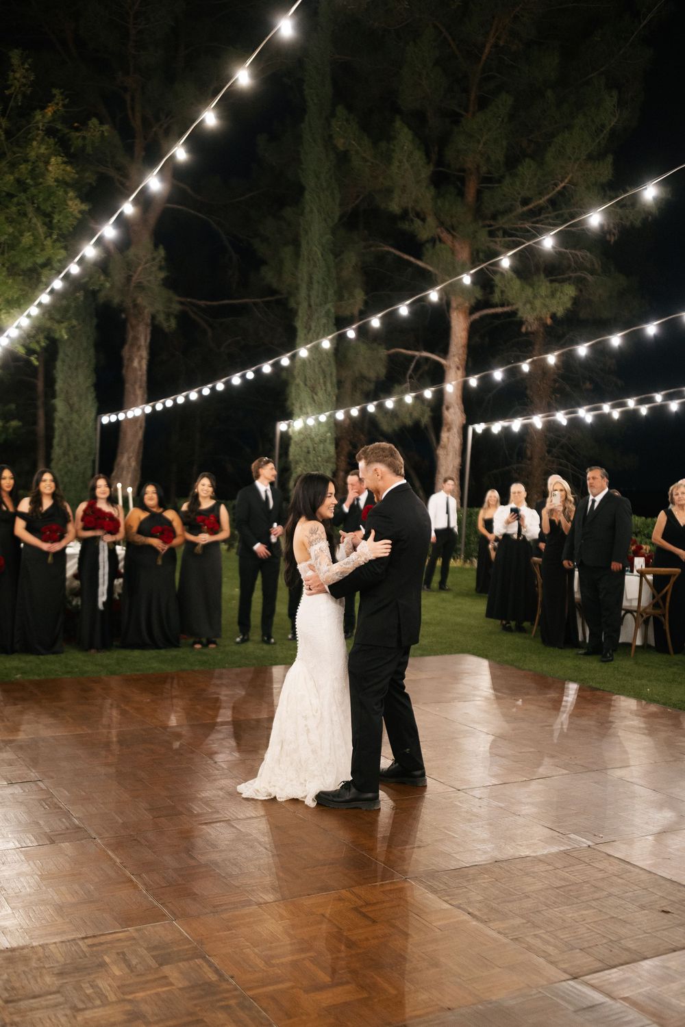 Bride and groom share first dance on outdoor wooden floor under string lights at elegant evening wedding reception.