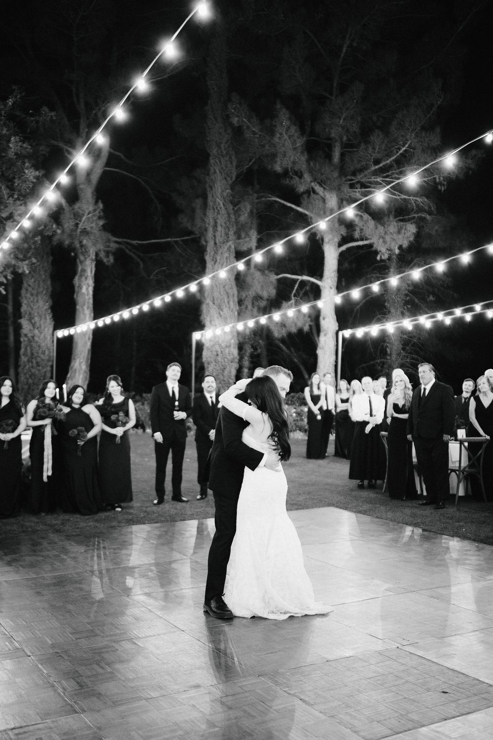 Bride and groom share first dance on outdoor wooden floor under string lights at night wedding reception.
