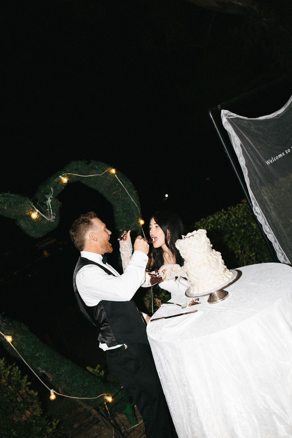 Bride and groom cutting their white wedding cake outdoors at night, surrounded by string lights and heart-shaped greenery.