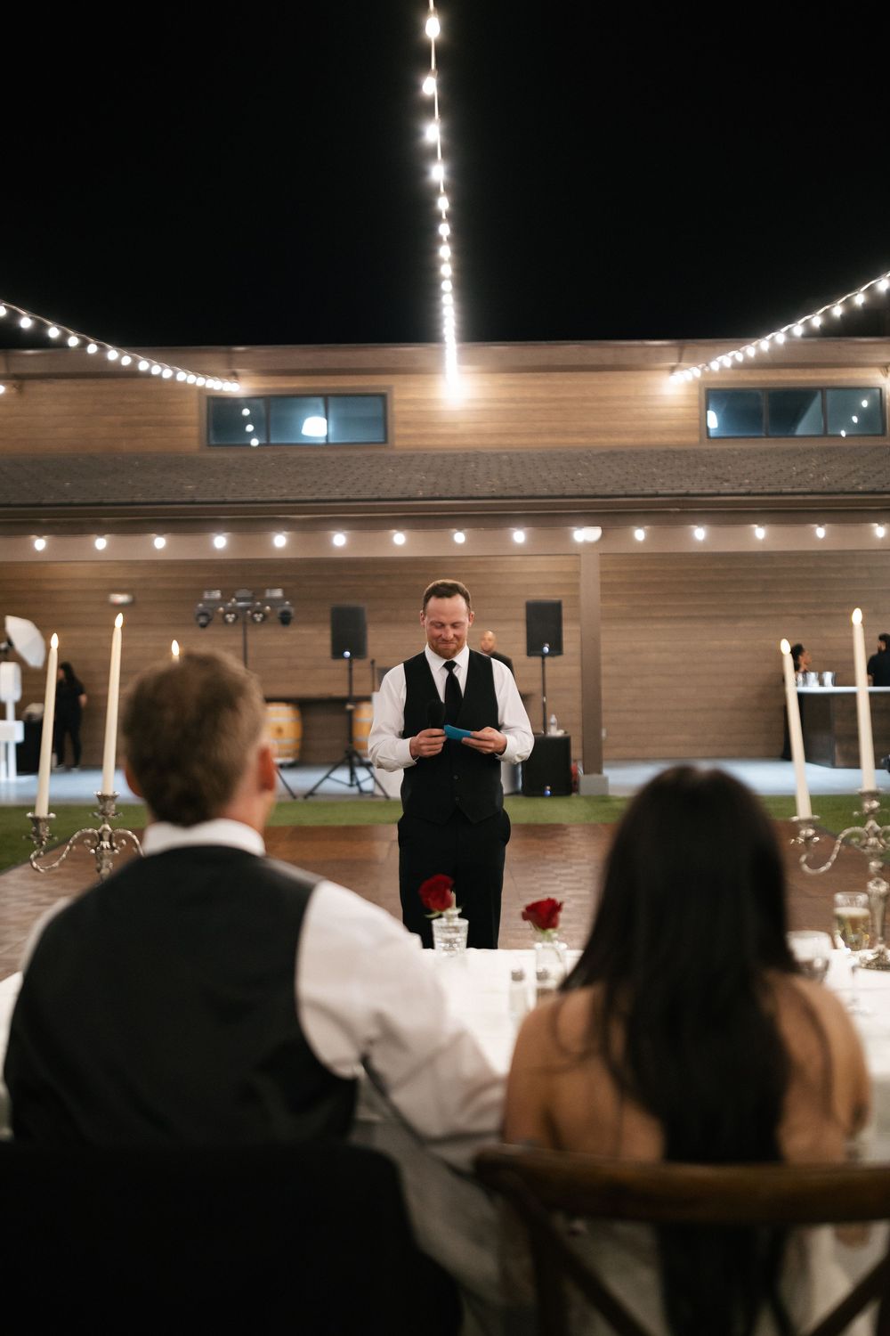 A man in a black vest delivers a heartfelt wedding speech outdoors at night under string lights to seated couple.