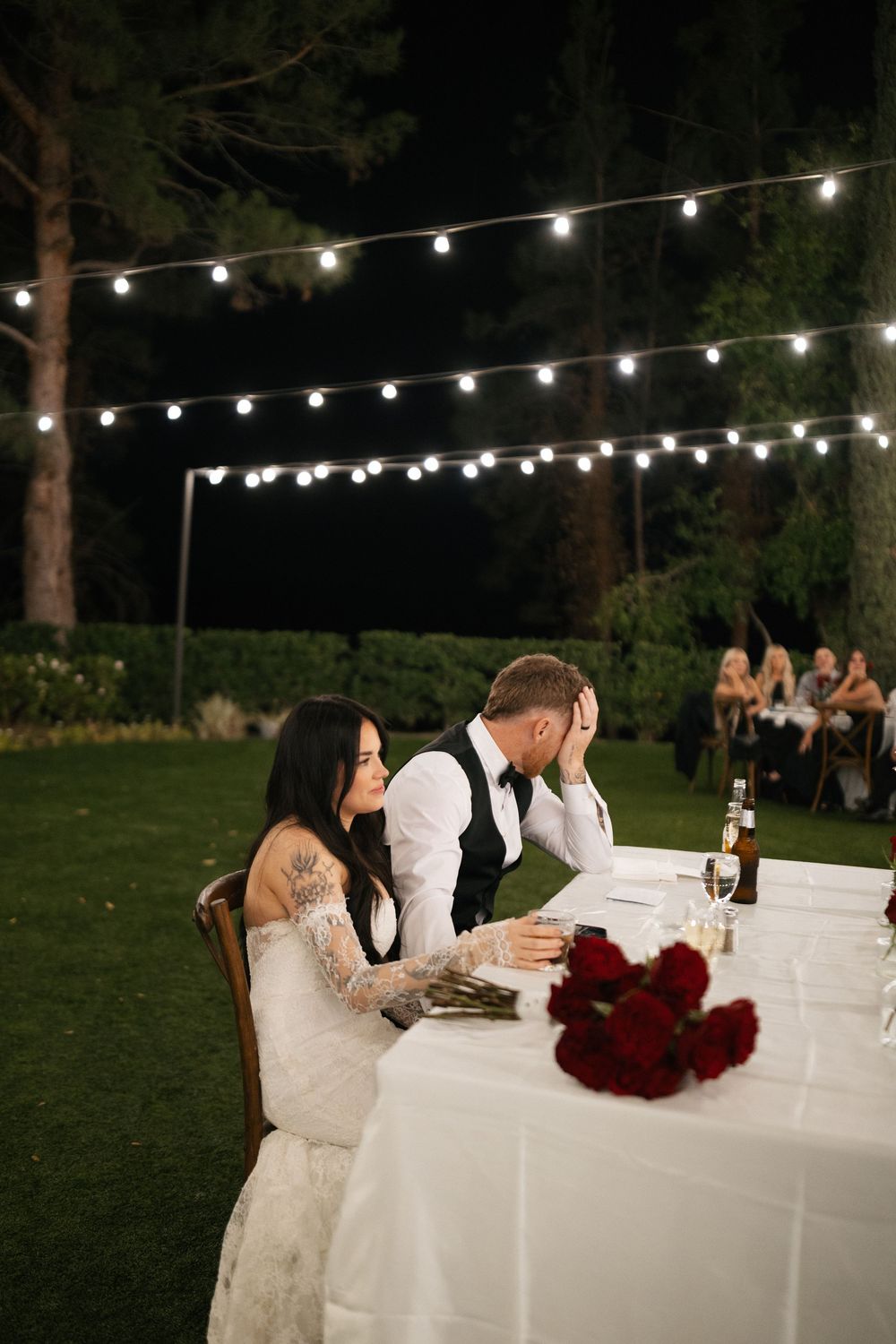 Bride and groom sit at sweetheart table during outdoor evening wedding reception with string lights and red roses.