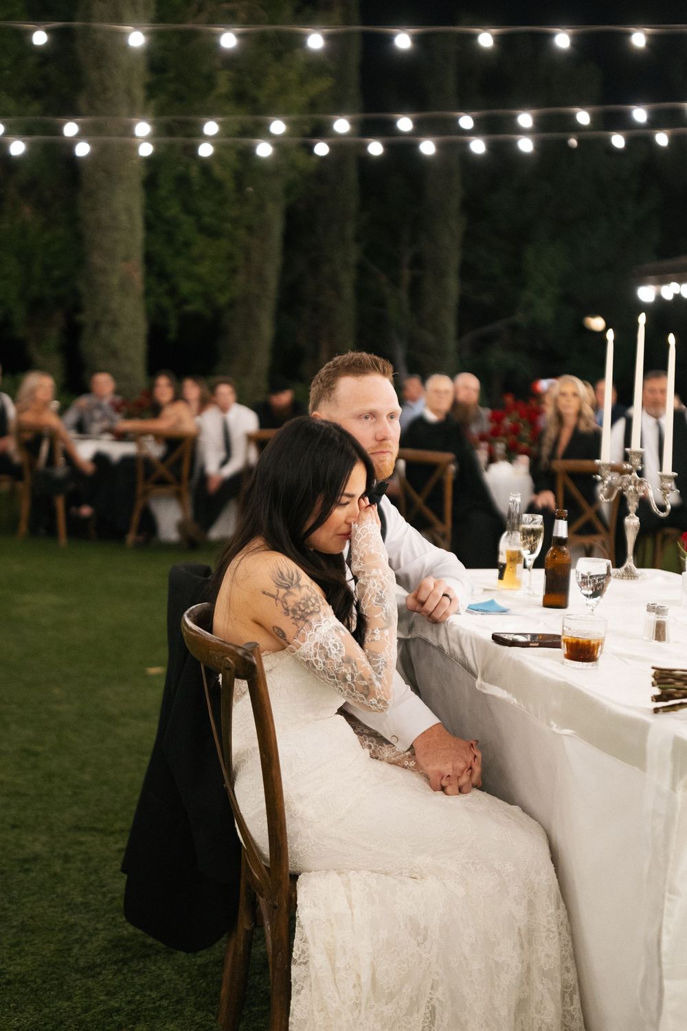 Bride and groom at outdoor wedding reception table, string lights glowing above guests seated on lawn. Micro wedding, elopement, intentional
