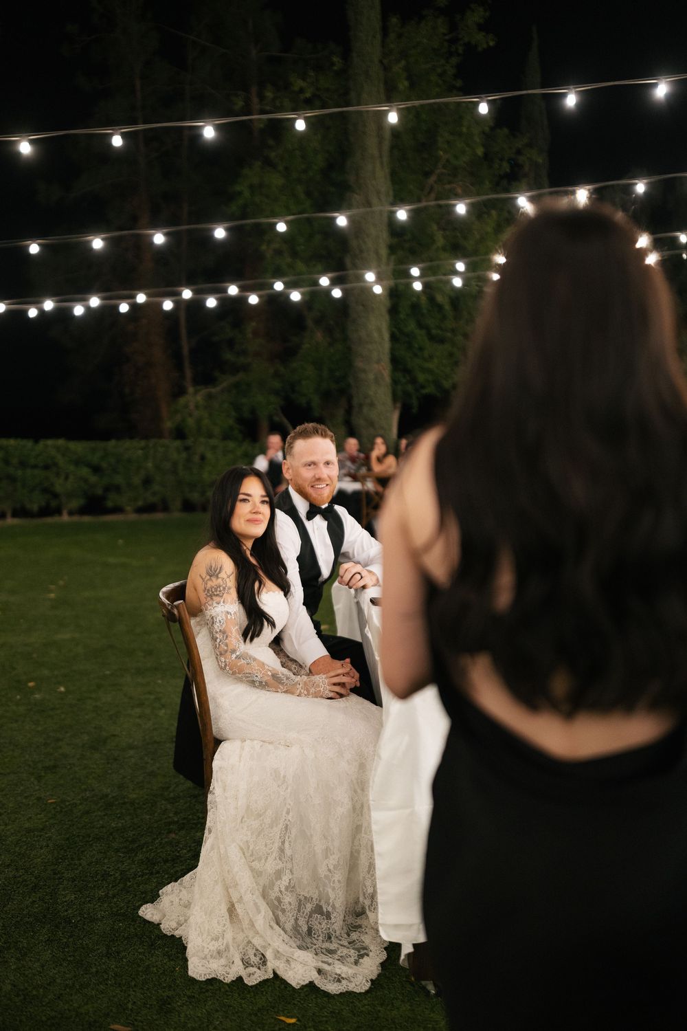 Bride in lace gown and groom in vest smile at guest during outdoor nighttime wedding reception with string lights.
