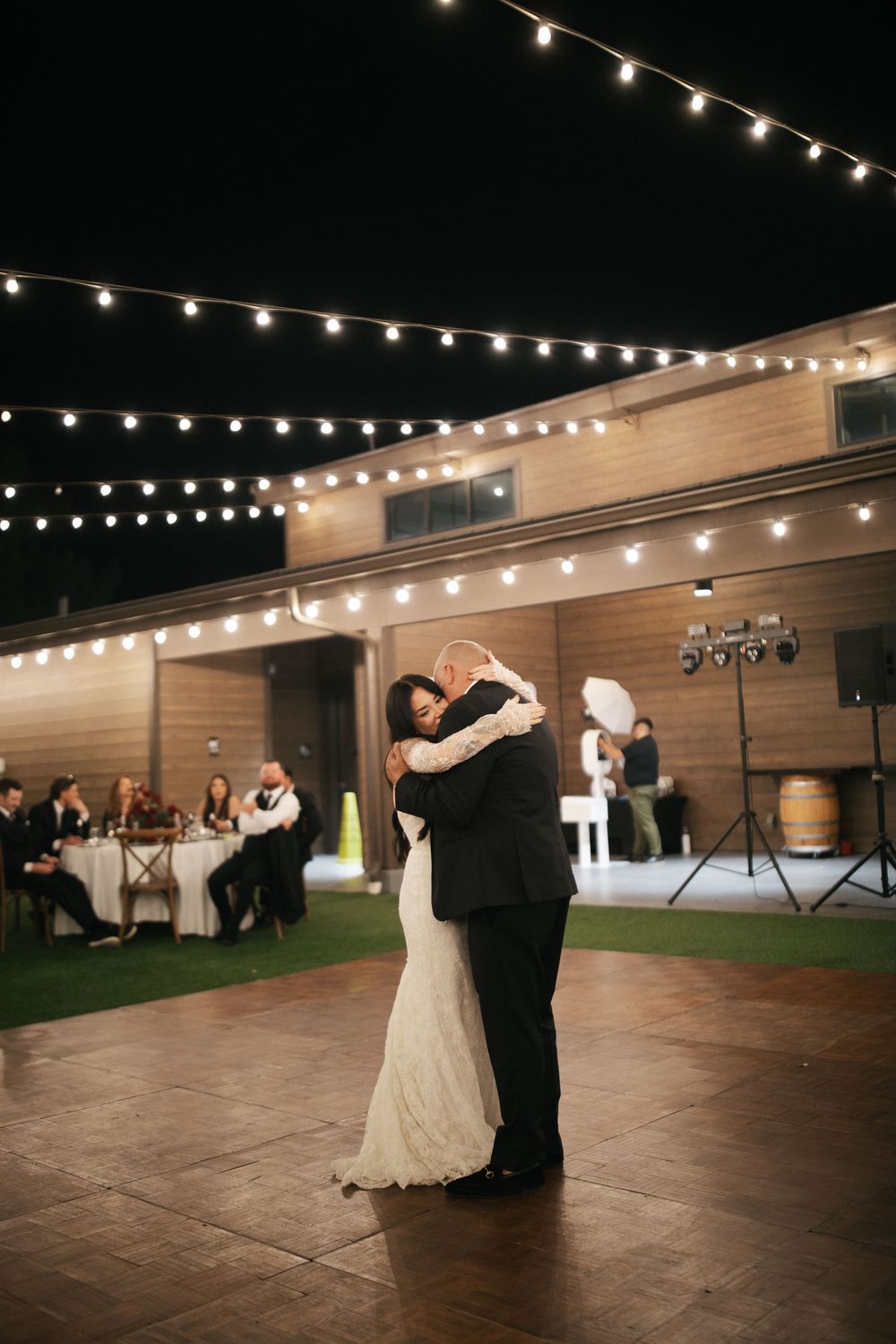 Bride and groom share their first dance under string lights at an outdoor wedding reception at night.