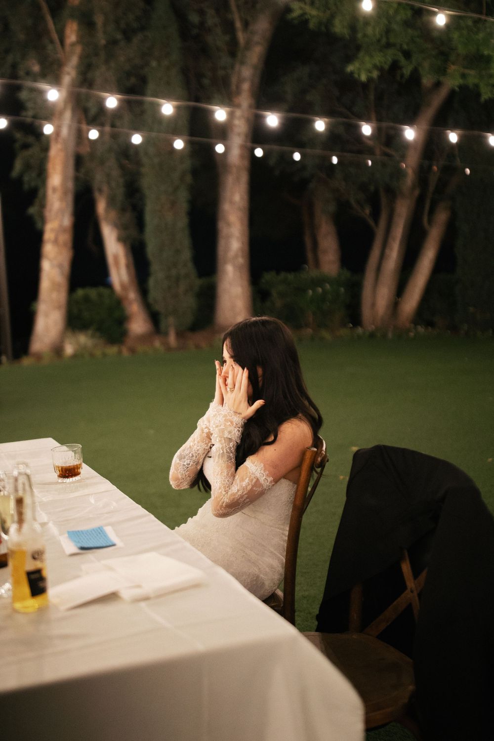 Emotional bride in lace dress covers her face at sweetheart table during outdoor evening wedding reception.