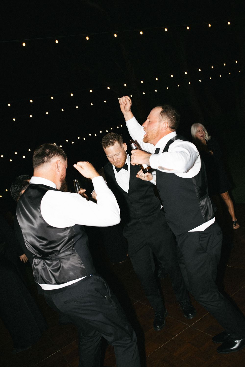 Groomsmen in black vests and bow ties dancing energetically at an outdoor wedding reception under string lights.