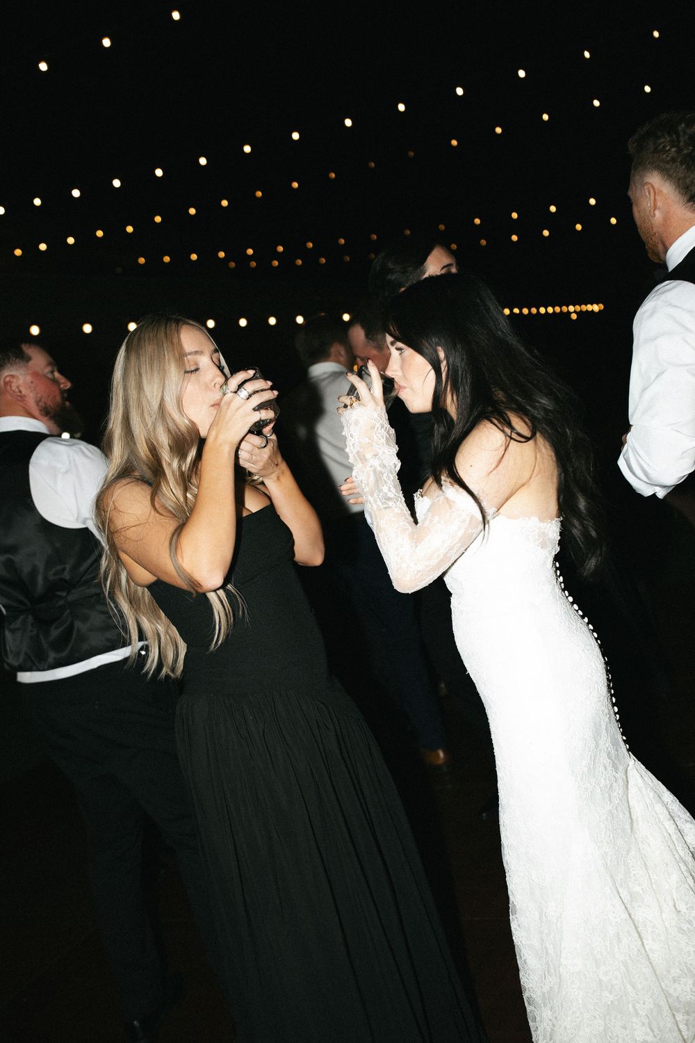 Bride in lace gown dancing with guest in black dress at wedding reception under string lights.