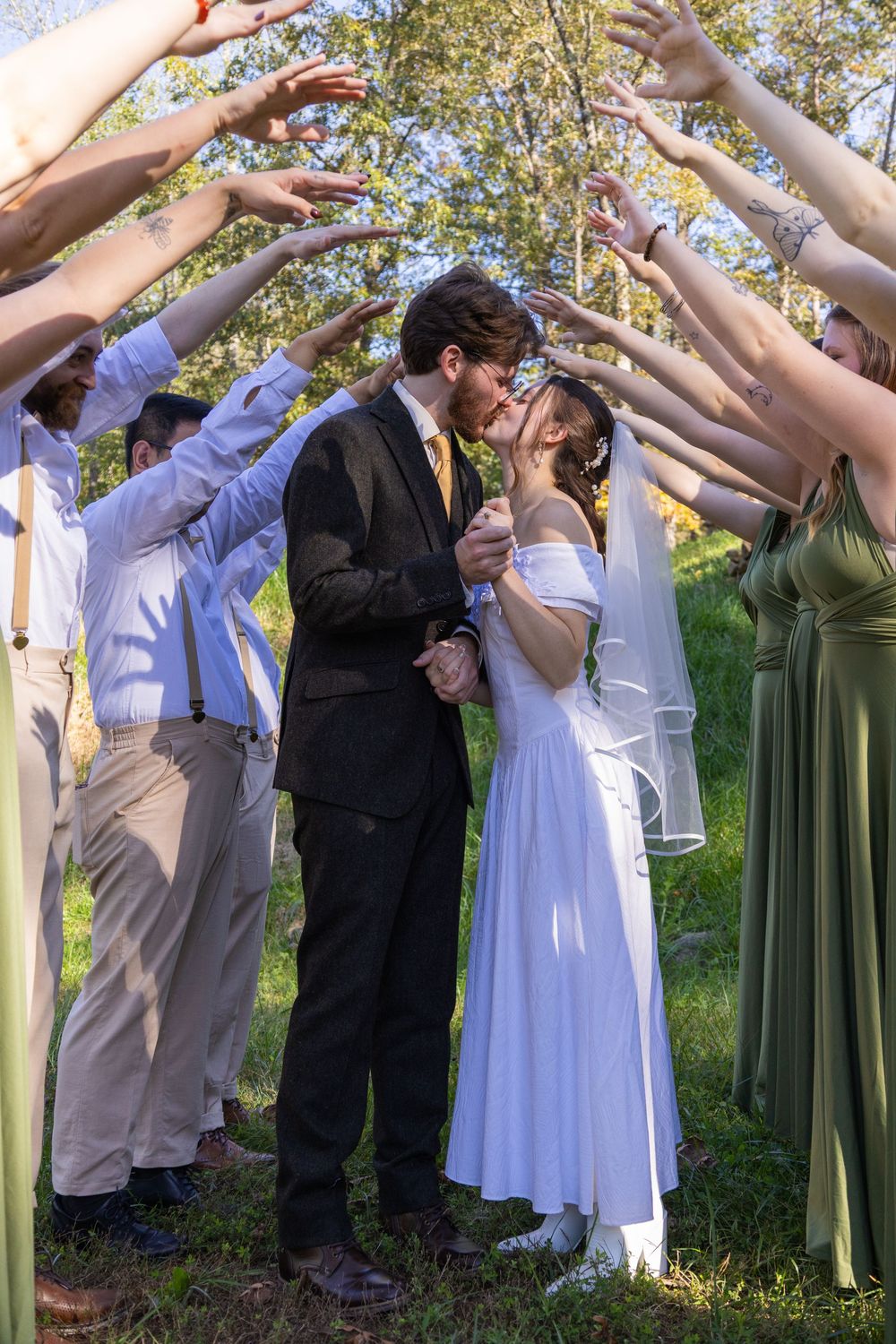 Wedding party forms an arch with their arms as bride and groom share a kiss outdoors.