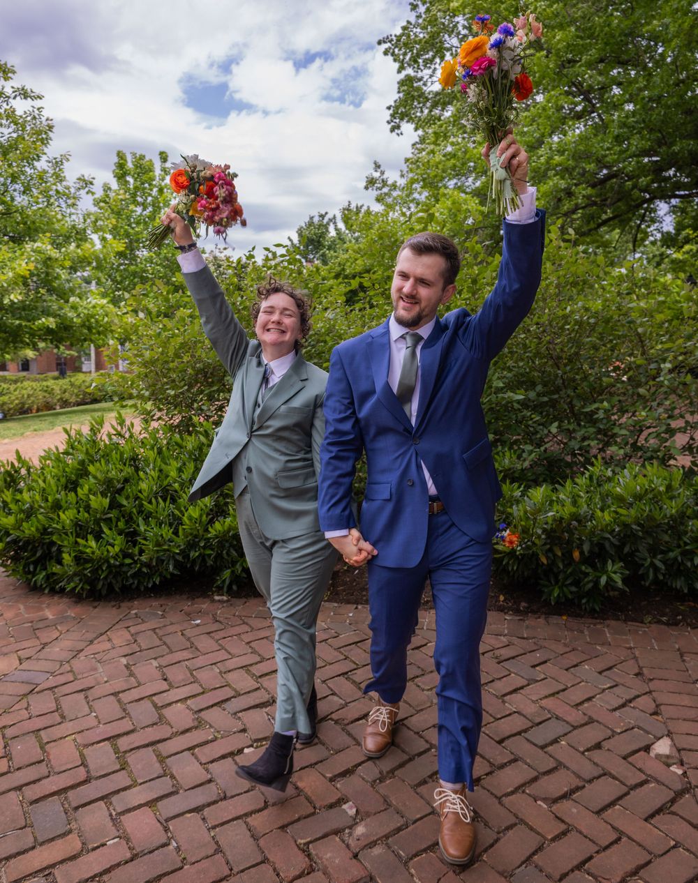Two newlyweds in suits celebrate joyfully, raising colorful bouquets on a brick path surrounded by lush green trees.