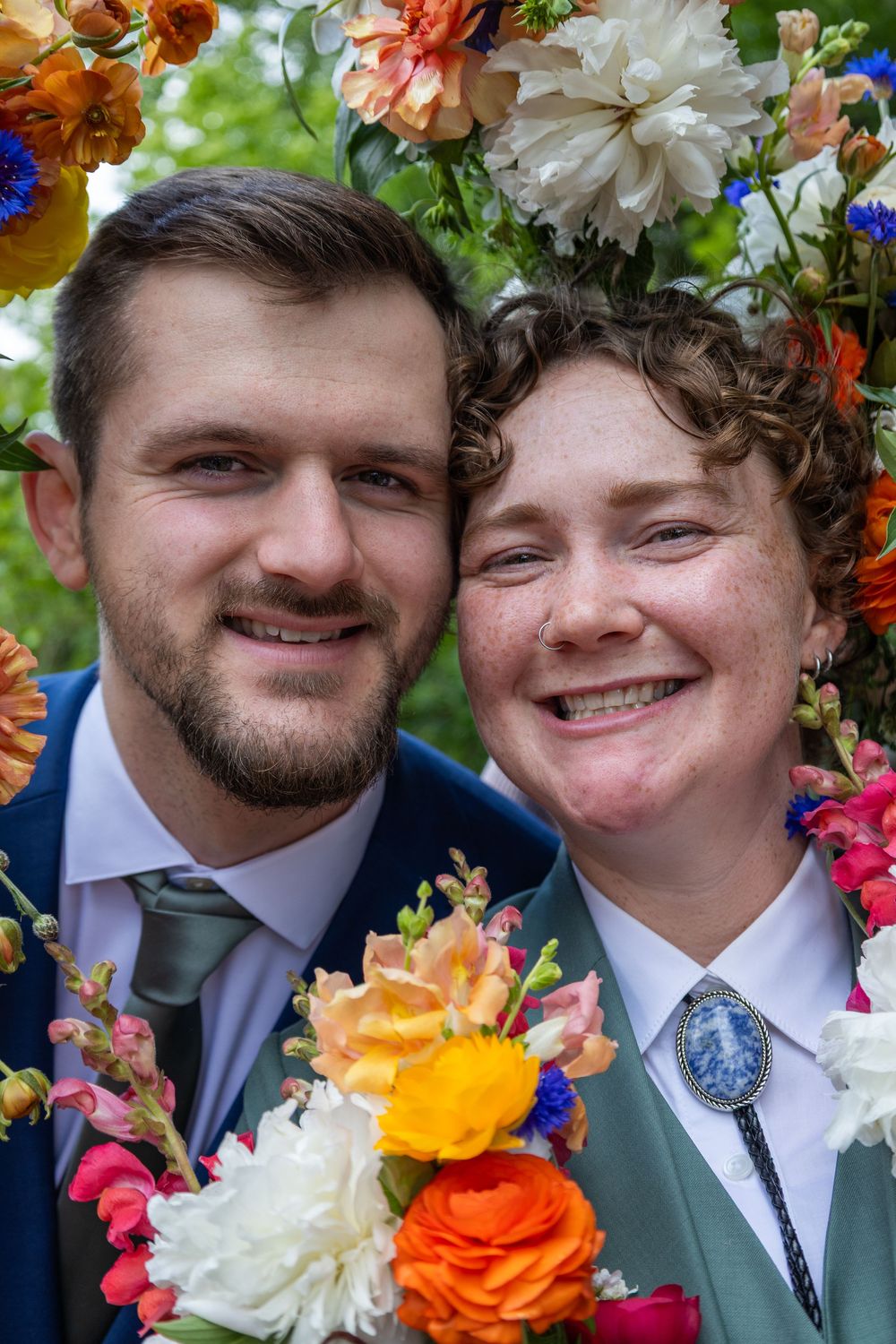 Smiling couple posing together under colorful floral arch, man in blue suit with tie, woman wearing bolo tie with brooch.