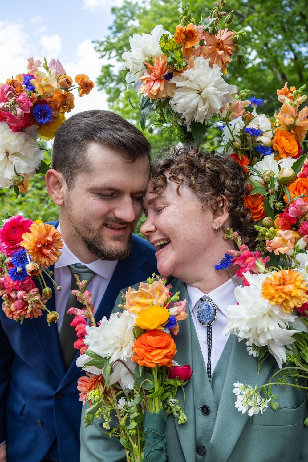 Couple in formal attire smiling together surrounded by vibrant colorful flowers including orange dahlias and pink blooms.