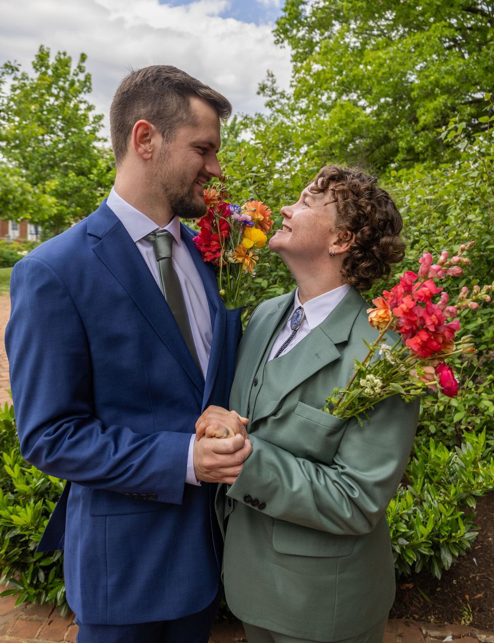 Two people in suits holding hands and smiling at each other outdoors, surrounded by colorful flowers.