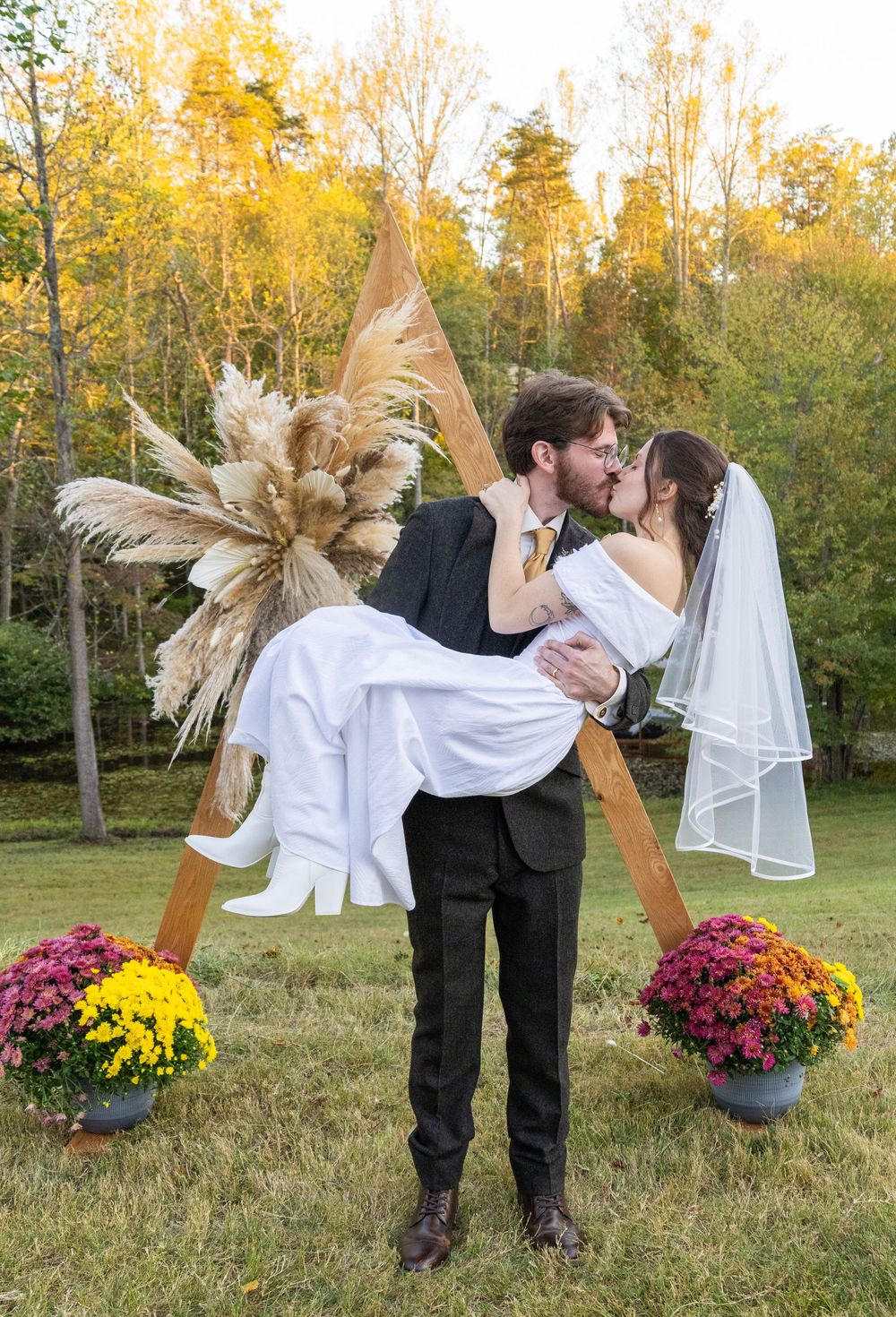Groom carries bride in his arms, kissing her in front of a wooden triangular wedding arch.