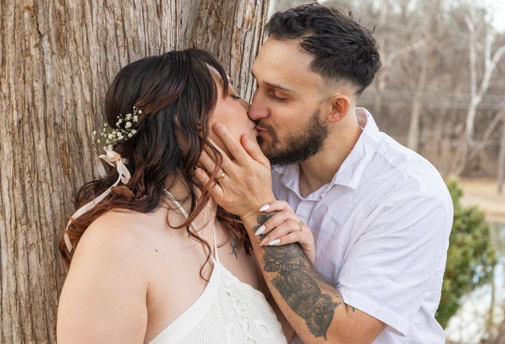 Bride and groom share a romantic kiss against a tree, with floral hair accessories and tattoos visible.
