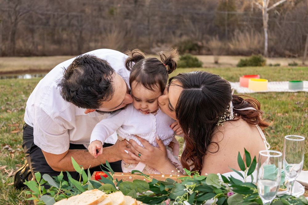 Family portrait with parents kissing baby on cheeks during outdoor picnic on green grass.