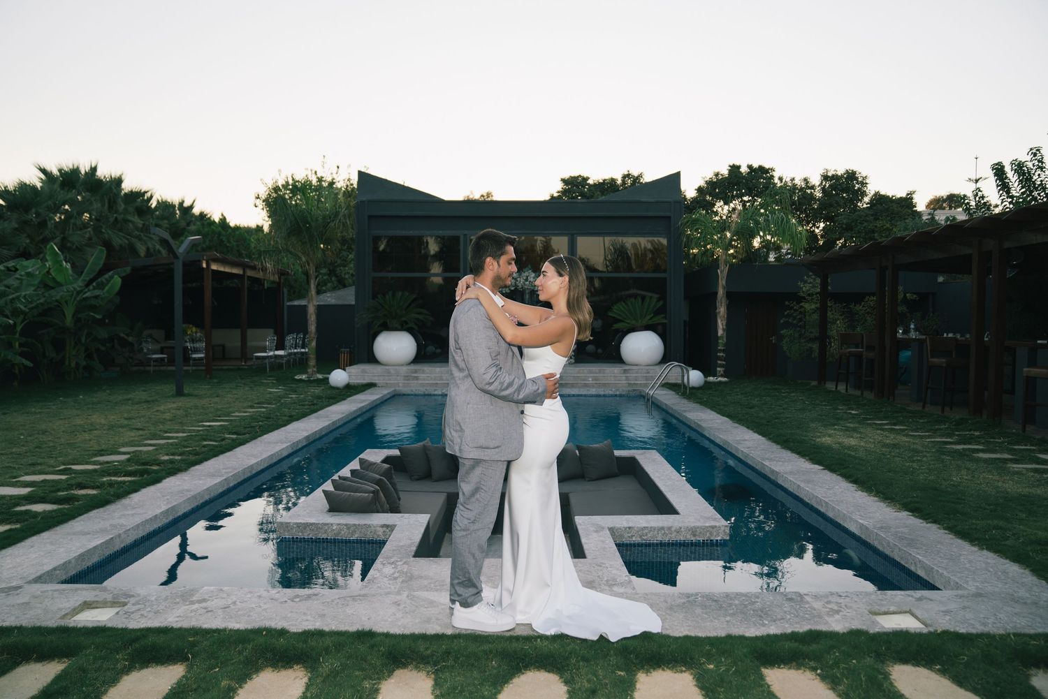 Bride and groom embrace romantically by an elegant pool at dusk during their outdoor wedding reception.