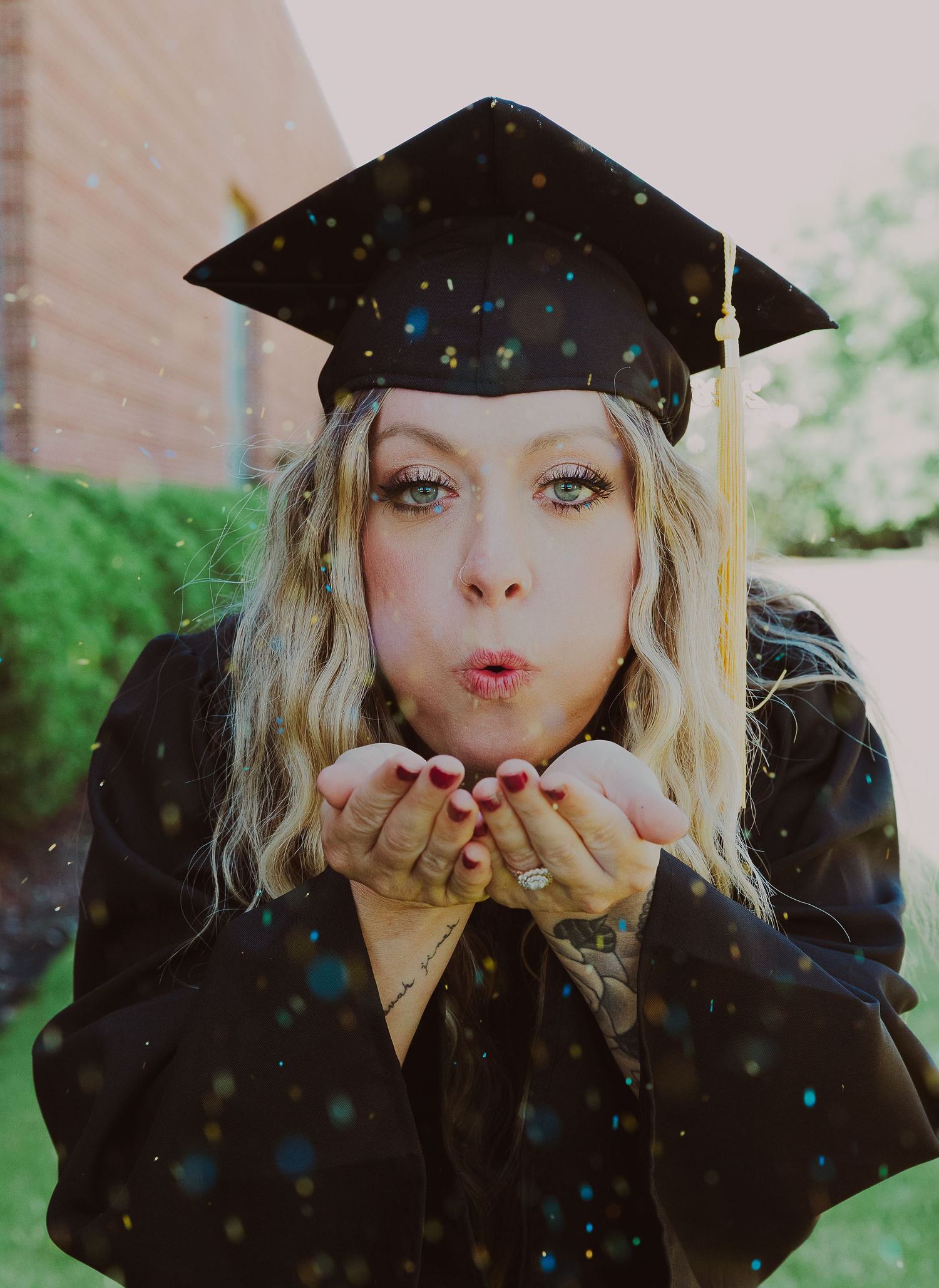 Graduate in black cap and gown blowing glitter from hands, celebrating graduation outdoors.