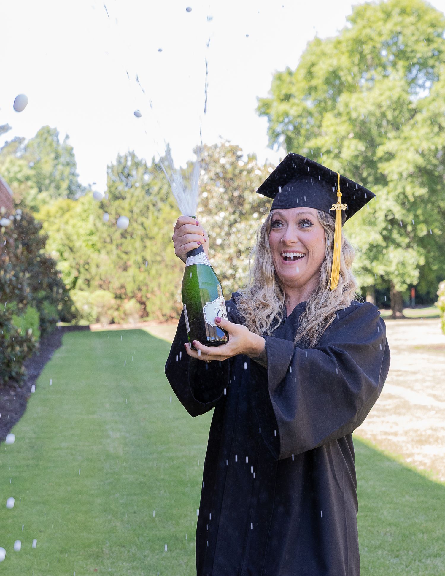 Woman in graduation cap and gown celebrates with popping champagne bottle outdoors on sunny day.