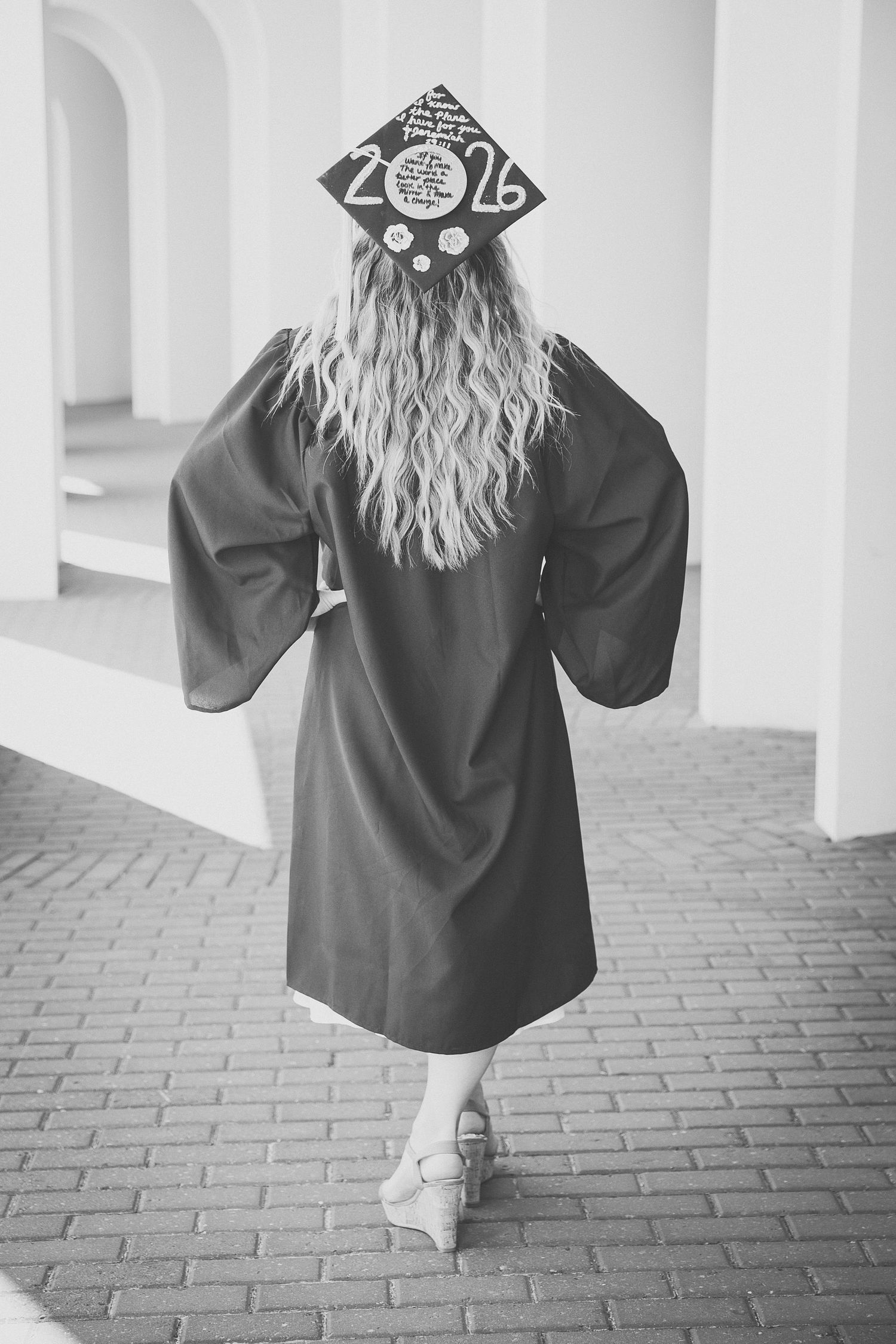 Black and white photo of a graduate in cap and gown with long blonde hair walking through arched columns.