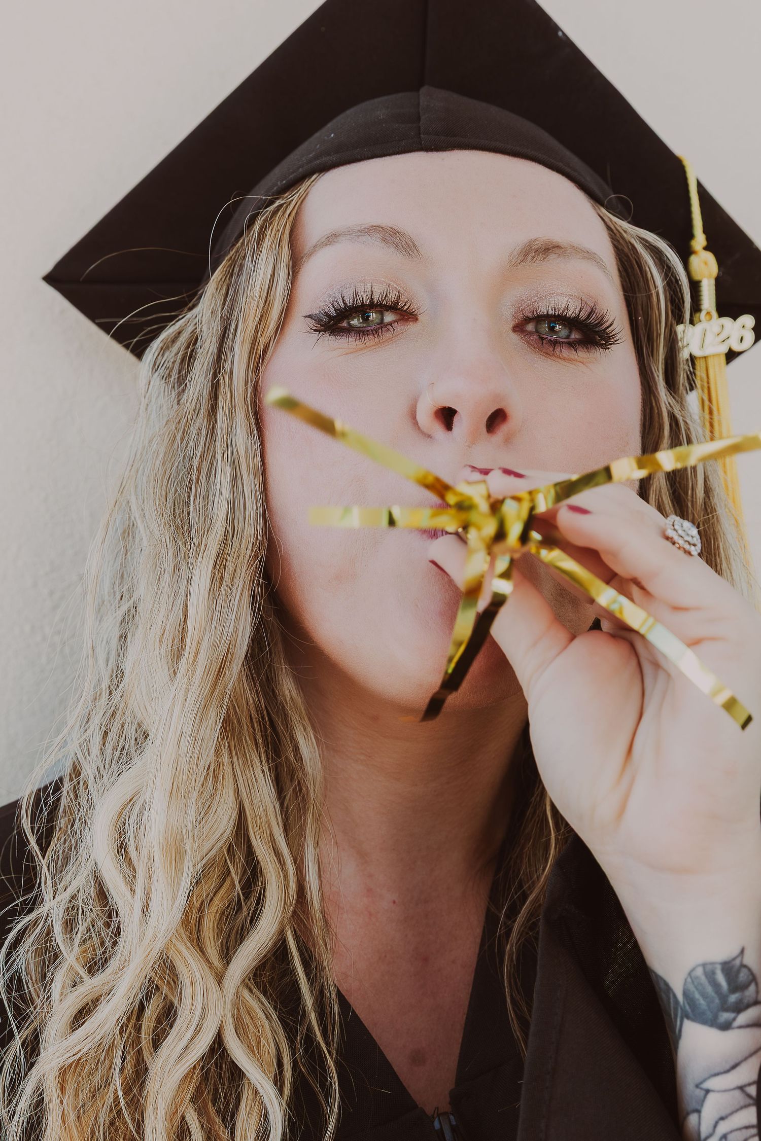 A blonde woman in a black graduation cap blows gold confetti streamers, celebrating her 2023 graduation milestone.