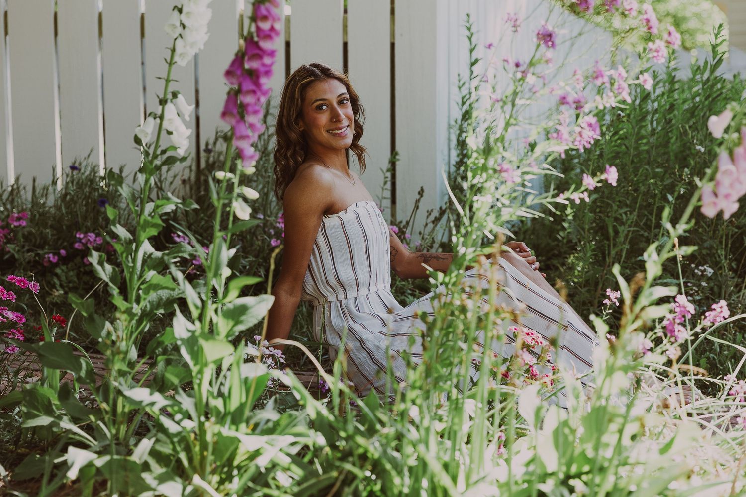 Young woman in striped dress sitting among colorful wildflowers beside a white fence in a lush garden.