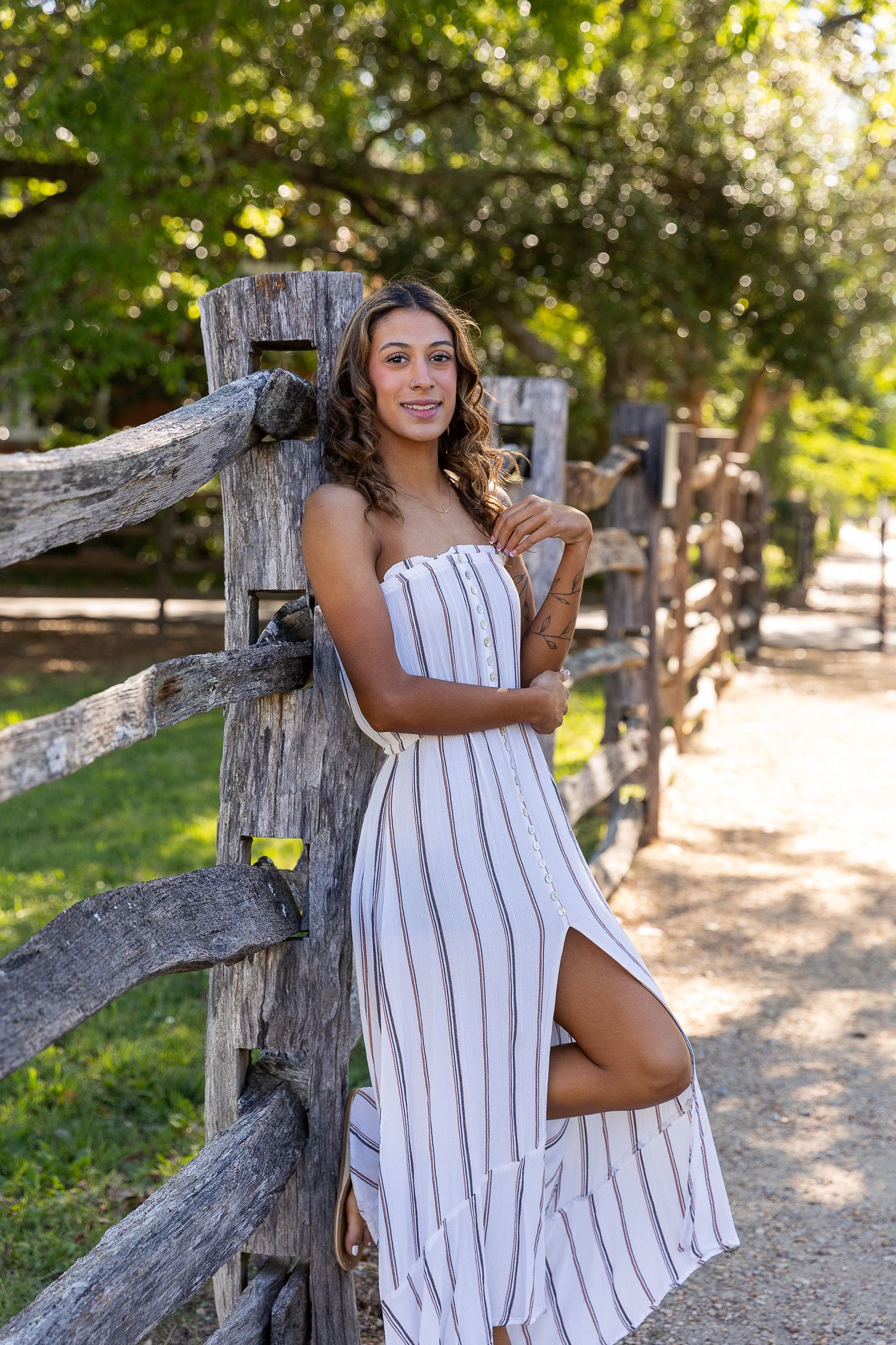 Young woman in striped white dress leaning against rustic wooden fence surrounded by lush green trees.