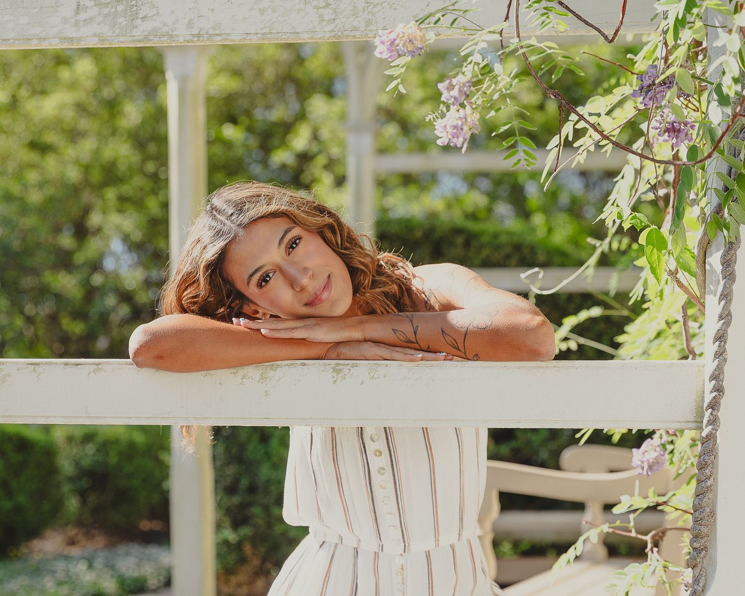 Young woman in white striped dress leaning on garden fence surrounded by wisteria and lush greenery.