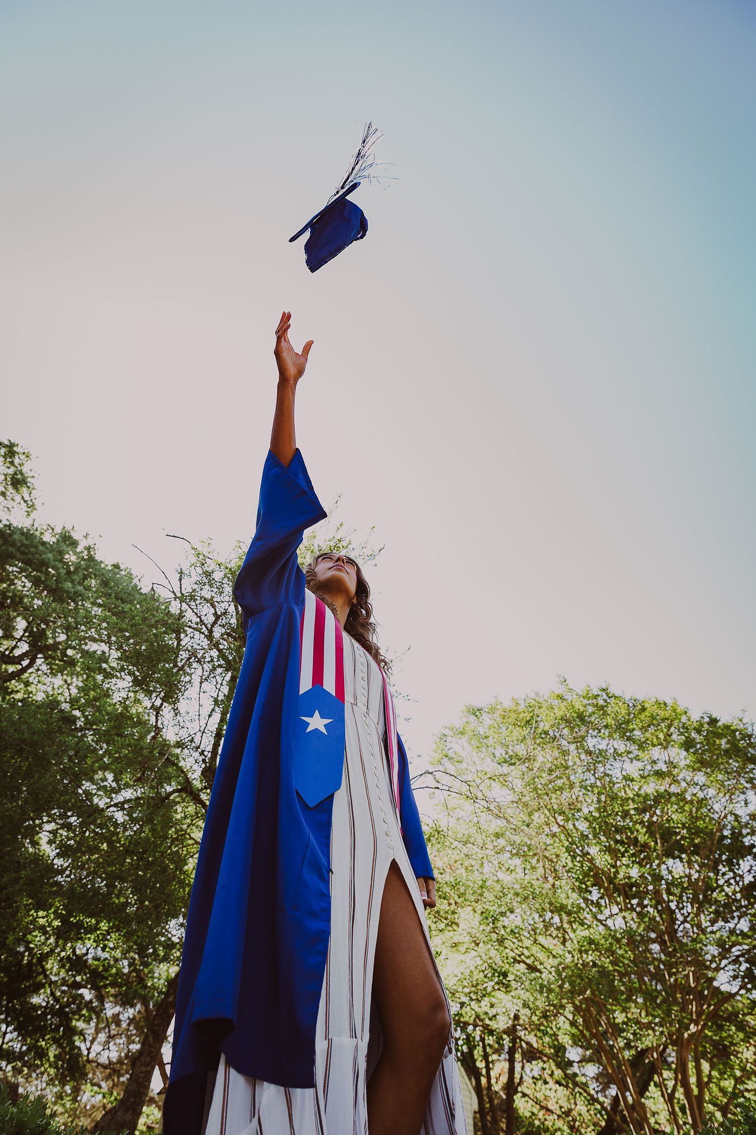 Graduate in blue cap and gown tossing mortarboard into sky celebrating graduation outdoors.
