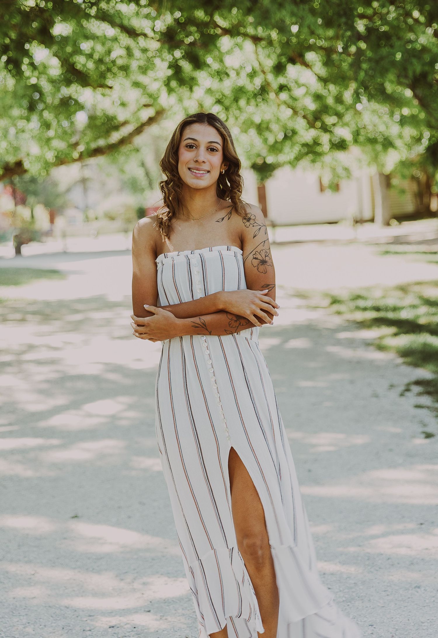 Young woman in striped maxi dress with slit stands arms crossed outdoors under green trees, smiling confidently.