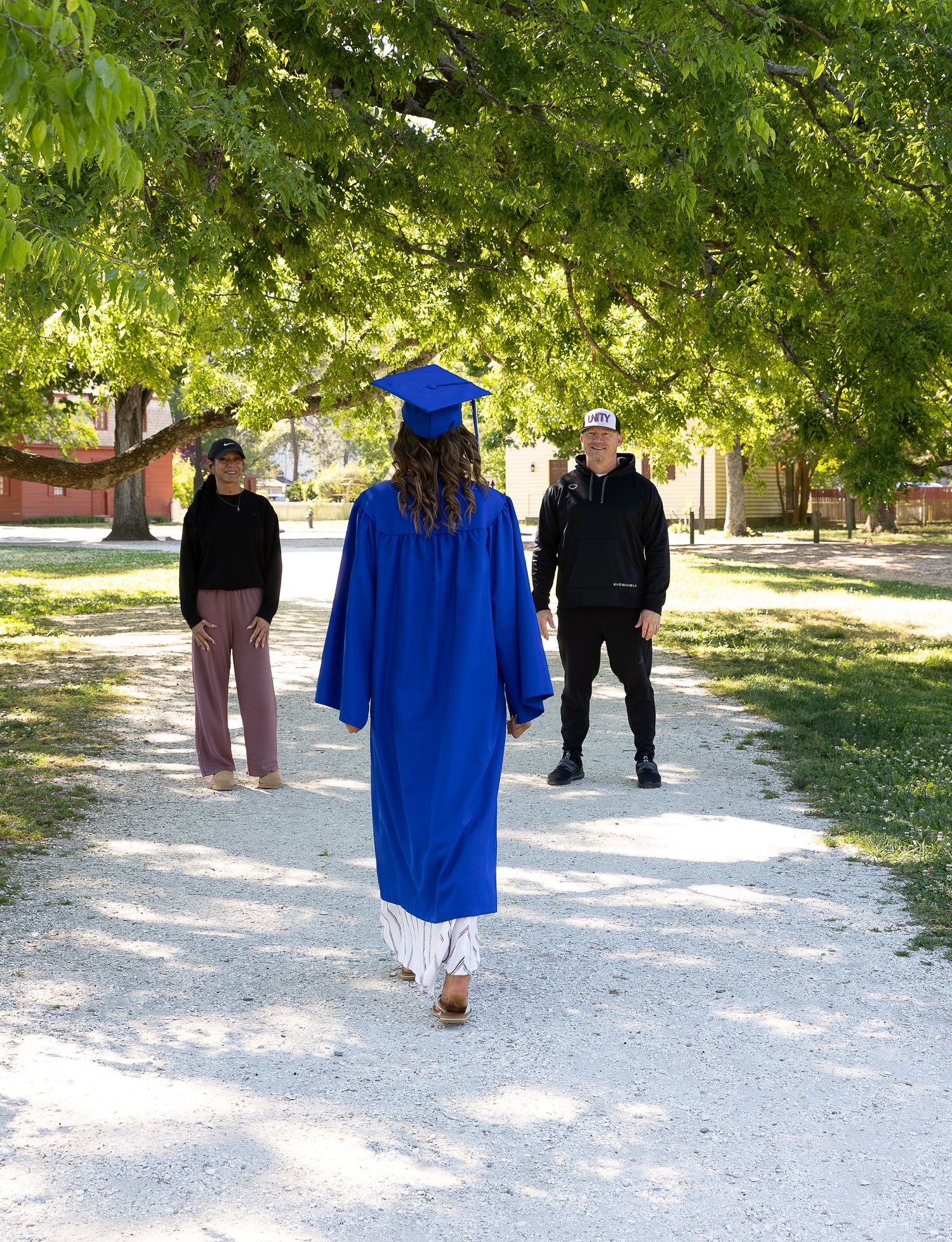 Graduate in blue cap and gown walks along a tree-lined path toward two people during an outdoor graduation photo session.