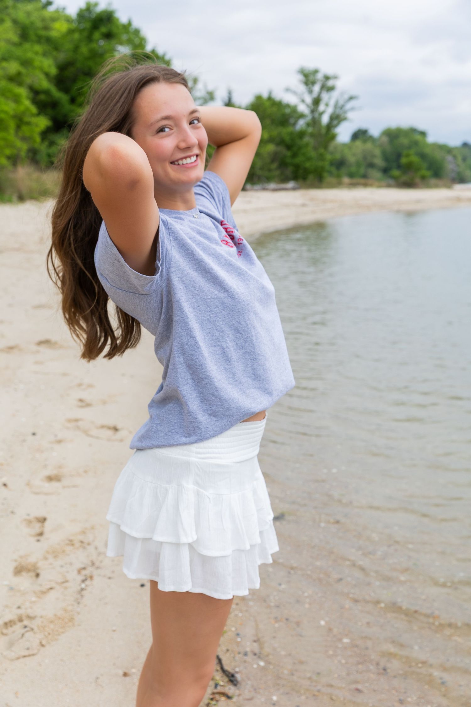 Smiling teenage girl in gray tee and white ruffle skirt posing on sandy beach shore.