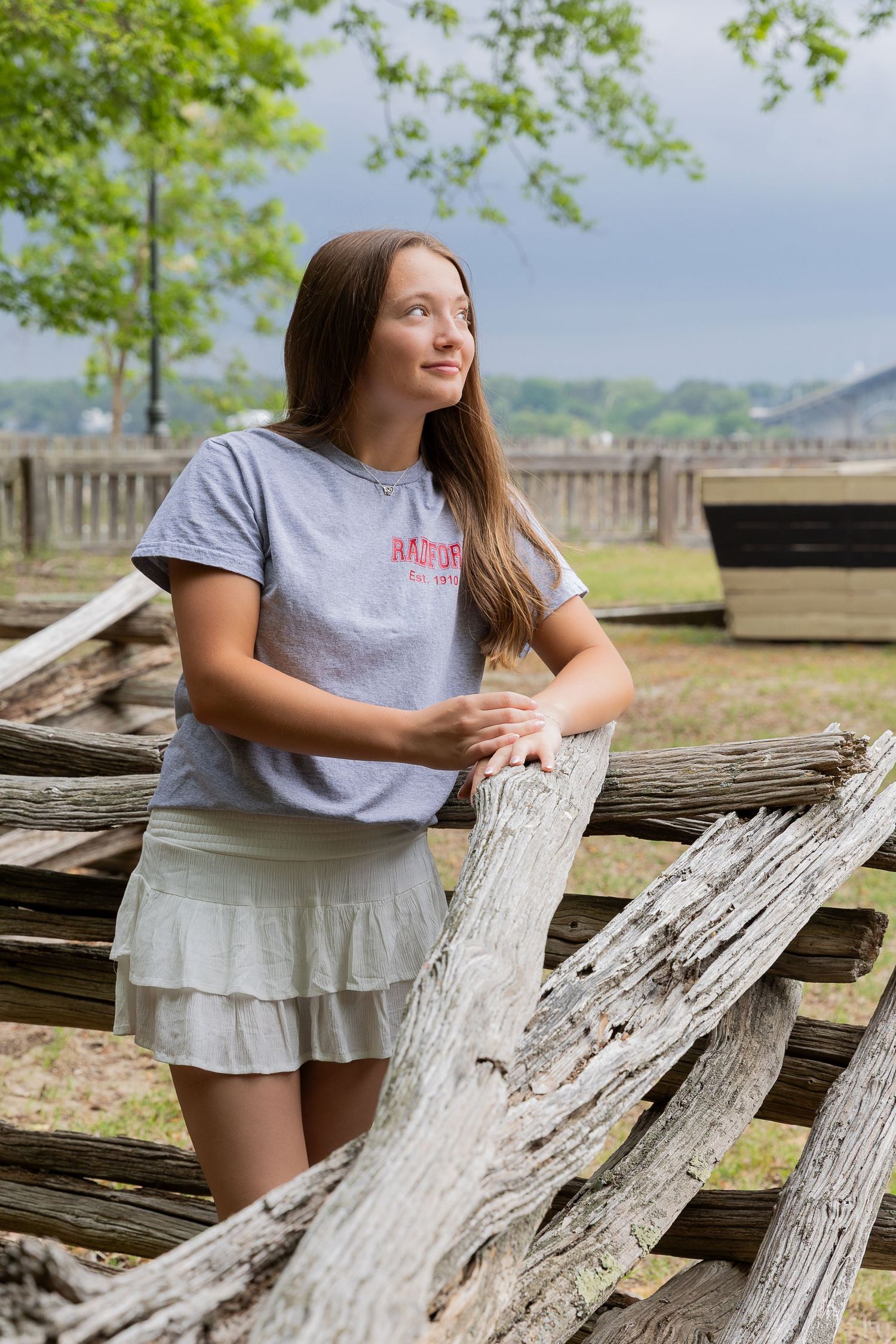 Teenage girl in gray tee and ruffle skirt leaning on rustic wooden fence outdoors.