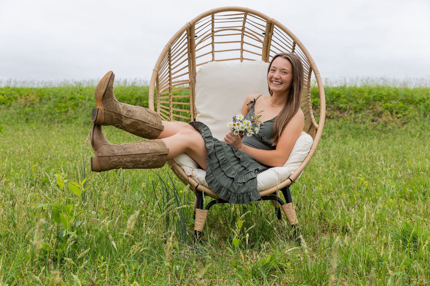 Young woman in cowboy boots and floral dress relaxing in a wicker egg chair on a green grassy field.