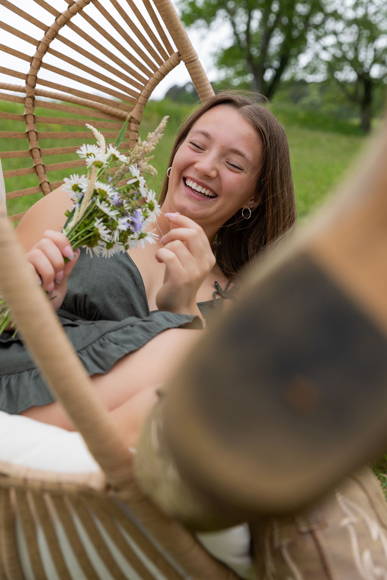Young woman laughing in a rattan chair outdoors, holding wildflowers, wearing a green dress.