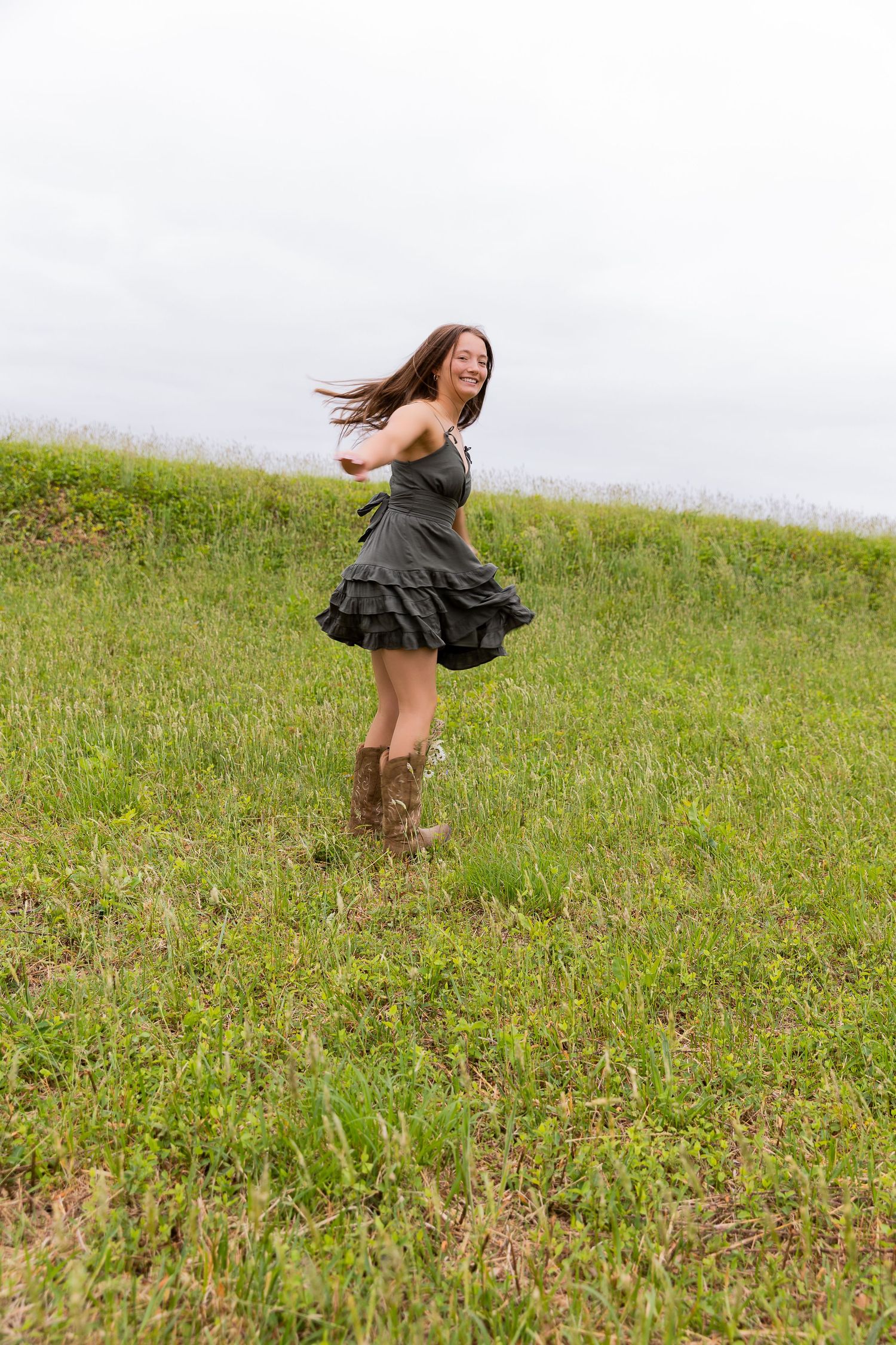 Young woman in gray ruffle dress and cowboy boots dancing joyfully on a green grassy hillside.