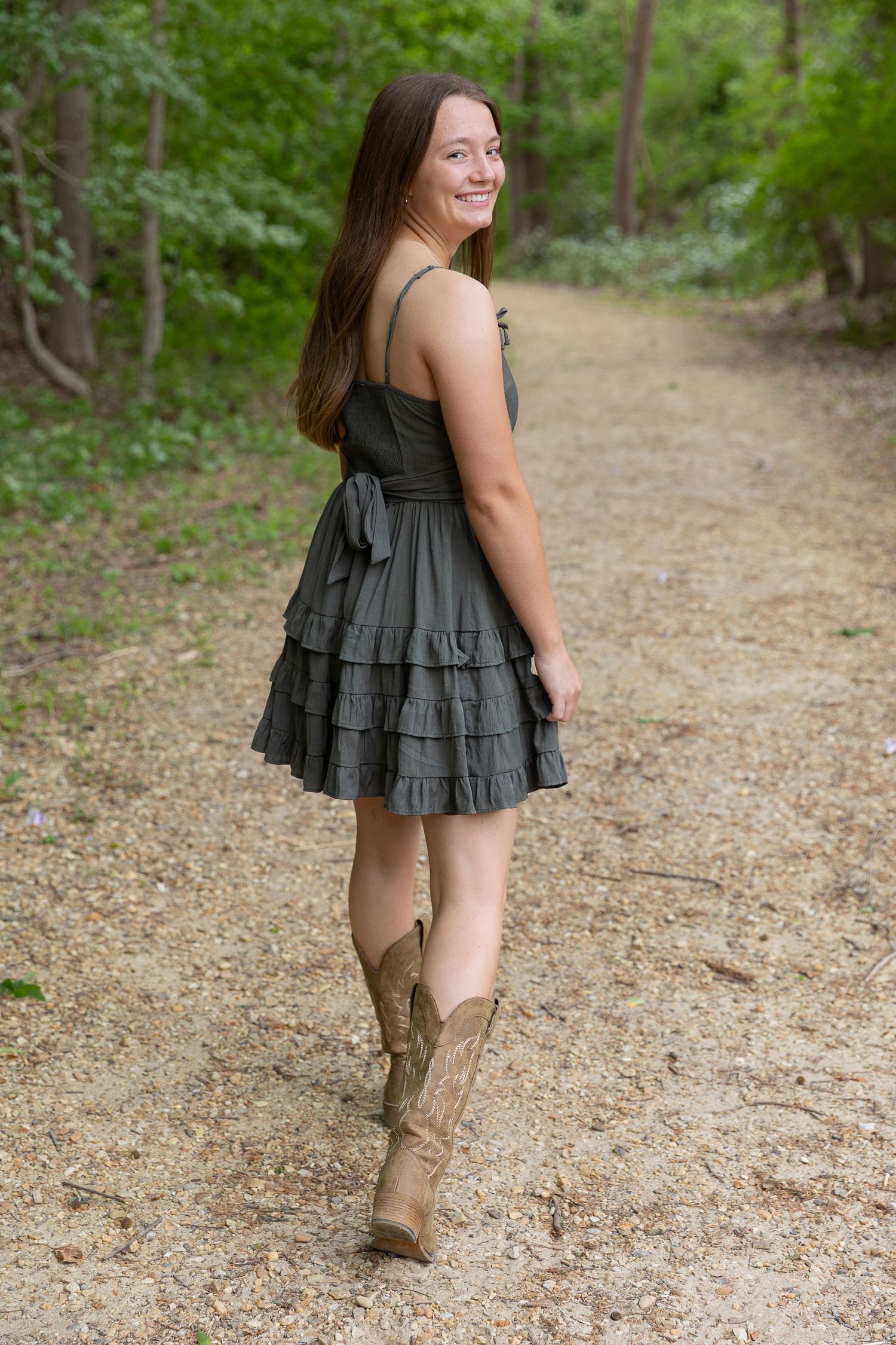 Smiling young woman in gray ruffle dress and cowboy boots walking on a wooded dirt path.